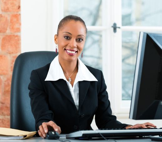 Mulher com largo sorriso, cabelos presos, sentadao em frente ao computador, segurando o mouse com a mão direita e apoiando a mão esquerda no teclado